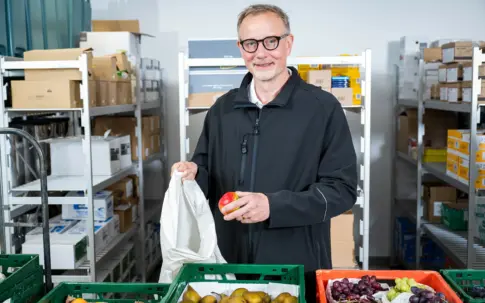 Mann hält Apfel und Stoffbeutel zwischen Kisten mit Obst in einem Lagerraum.
